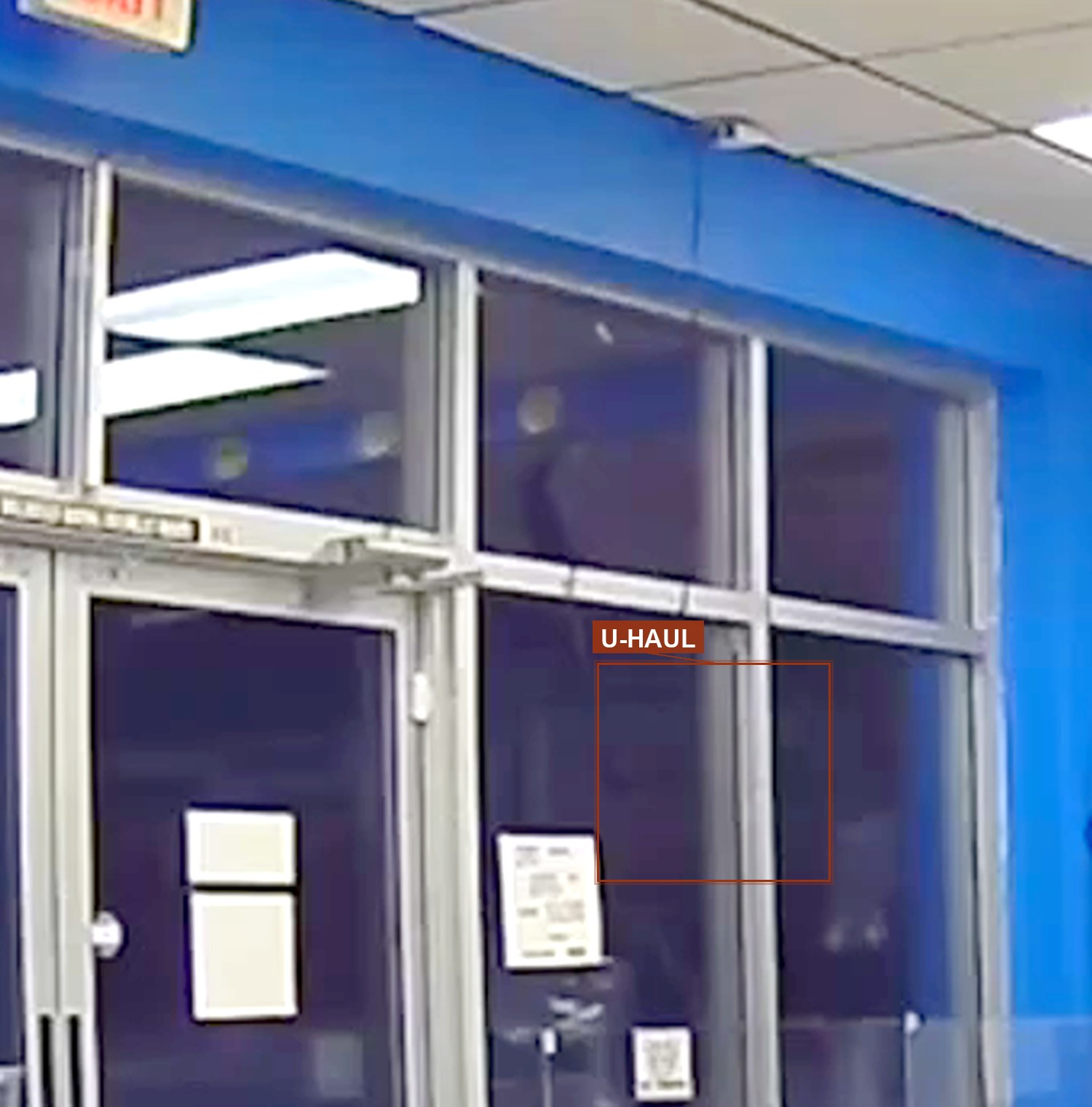 A frame from the store's own security camera looking out through the blue storefront glass at night. In the lower-right pane, marked, a U-Haul truck is parked at the curb outside.