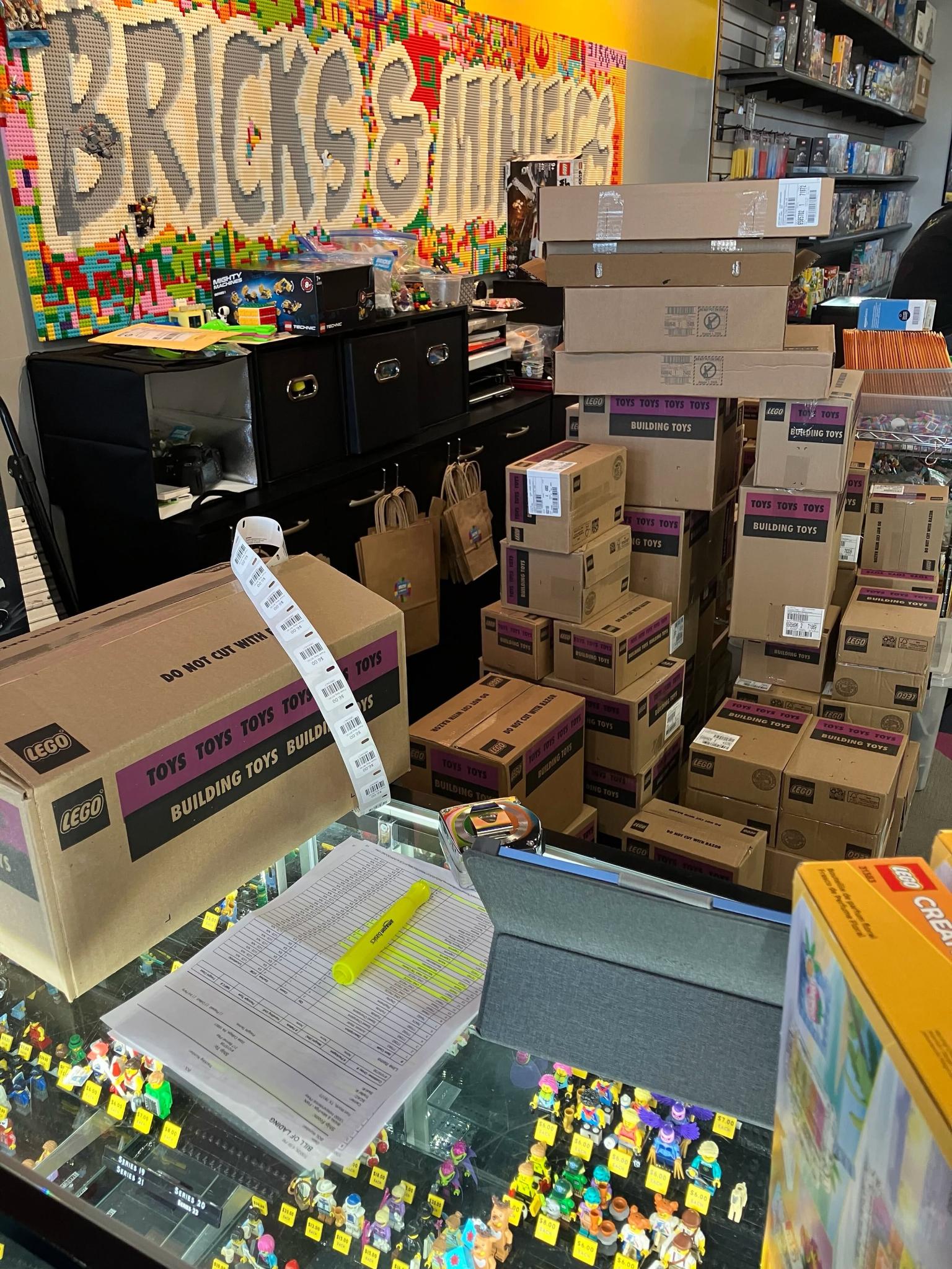A bill of lading on the glass front counter of a Bricks and Minifigs store, with stacks of sealed LEGO master cartons behind it and the store’s brick-built sign on the wall.