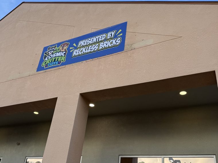 The same gable with the channel letters gone. A blue vinyl banner reading Cosmic Critter Toys, presented by Reckless Bricks, hangs across the rectangular outline the old sign left in the stucco.