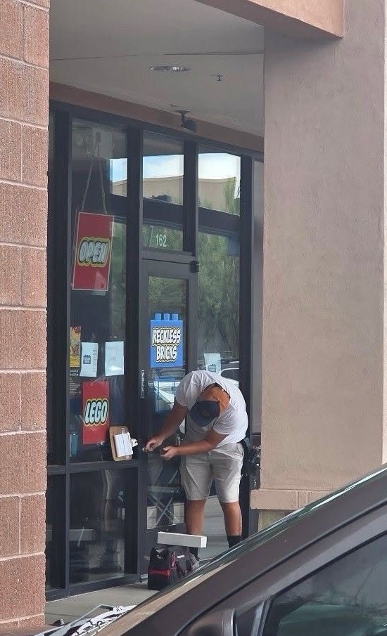A man in a white shirt and cap bent over the lock of a glass storefront door marked 162, a tool bag at his feet and a clipboard hung on the pull handle. In the window behind him: a blue Reckless Bricks sign, an OPEN sign and a LEGO sign.